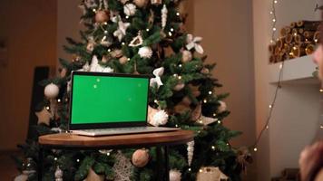 frau kommuniziert über einen videoanruf mit einem laptop mit grünem bildschirm und einer farbigen taste und feiert das neue jahr vor dem hintergrund des weihnachtsbaums. Fernkommunikationskonzept. Soziale Distanz.