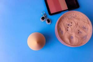 Beauty box, loose matte mineral powder and blush with a beauty blender for makeup and silver earrings on a blue background. Flat lay. Top view photo