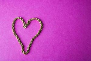 Texture of a beautiful golden festive chain unique weaving in the shape of a heart on a pink purple background and copy space. Concept love, marriage proposal, marriage, St. Valentine's Day photo