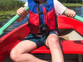 A man in a red boat, shorts and a life jacket is paddling with oars on a boat for a walk on the water of the lake river sea in nature photo