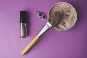 Beauty box, crumbly matte mineral powder with a special natural brown wooden brush from natural nap for make-up, pink perfume and shiny silver earrings on a pink purple background. Flat lay. Top view photo