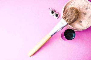 Beauty box, crumbly matte mineral powder with a special natural brown wooden brush from natural nap for make-up, pink perfume and shiny silver earrings on a pink purple background. Flat lay. Top view photo