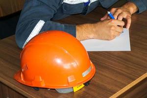 un hombre que trabaja como ingeniero con un casco amarillo naranja sobre la mesa está estudiando, escribiendo en un cuaderno en una fábrica de plantas industriales foto