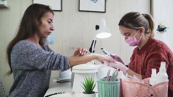 garota nos técnicos de unhas rosa uma máscara médica e luvas fazendo manicure para um cliente no salão de beleza. os cuidados com as unhas. video