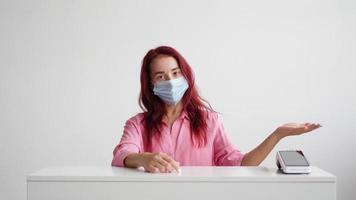 Woman administrator of a sales area in a pink shirt and a medical mask on her face stands behind the reception and invites clients to the store on a white background. Small business concept. video