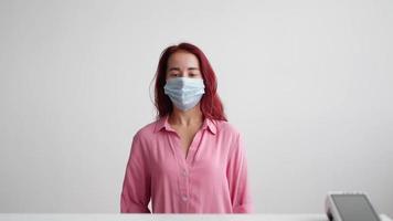 Woman administrator of a sales area in a pink shirt and a medical mask on her face stands behind the reception and invites clients to the store on a white background. Small business concept. video