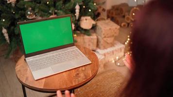 Woman communicates on a video call using a laptop with a green screen and a colored key, celebrating the new year against the background Christmas tree. Remote communication concept. Social Distance.