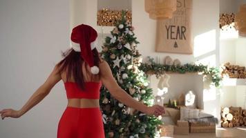 contento donna nel un' Santa Claus cappello corre per il Natale albero nel un' spazioso luminosa appartamento. nuovo anno, festivo umore. lento movimento. video
