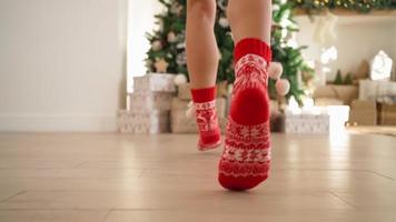 detailopname, gelukkig vrouw in rood nieuw jaar sokken loopt door de licht kamer naar de versierd Kerstmis boom met geschenken. nieuw jaar, feestelijk humeur. onherkenbaar persoon. langzaam beweging. video