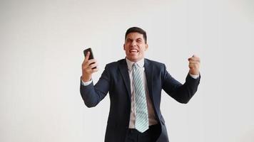 heureux employé de bureau masculin en vêtements formels utilise un téléphone portable et se réjouit de la victoire sur fond blanc. homme d'affaires en veste et cravate fait l'expérience du triomphe du gagnant, rit de joie. video