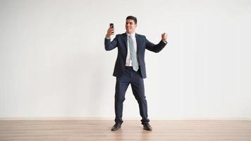 Happy male office worker in formal clothes uses mobile phone and rejoices in victory on white background. Businessman in jacket and tie experiences triumph of the winner, laughs with joy. video