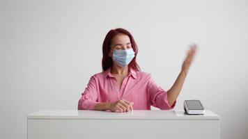 Woman administrator of a sales area in a pink shirt and a medical mask on her face stands behind the reception and invites clients to the store on a white background. Small business concept. video