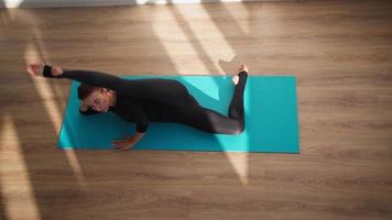 Athletic woman in sports overalls doing yoga and stretching while lying on a fitness mat at home on a wooden floor in bright sunlight. Top view. Slow motion. video