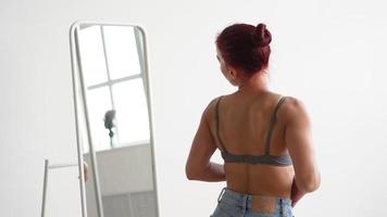 slank gelukkig vrouw in tank top en jeans loopt omhoog spiegel kijken Bij haarzelf. aantrekkelijk meisje looks in spiegel kiezen wat slijtage Bij huis Aan wit achtergrond. glimlacht en geniet zichzelf genieten van mooi zo ochtend. video