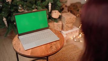 Woman communicates on a video call using a laptop with a green screen and a colored key, celebrating the new year against the background Christmas tree. Remote communication concept. Social Distance.