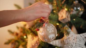 primer plano, una mujer irreconocible decora un árbol de navidad con bolas festivas. luces cálidas de guirnaldas parpadean en el fondo. vacaciones año nuevo. ambiente invernal, concepto de tradición. video