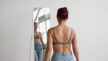 una mujer delgada y feliz con camiseta sin mangas y jeans corre hacia el espejo para mirarse a sí misma. chica atractiva se mira en el espejo eligiendo qué usar en casa con fondo blanco. sonríe y se divierte disfrutando de buenos días. video