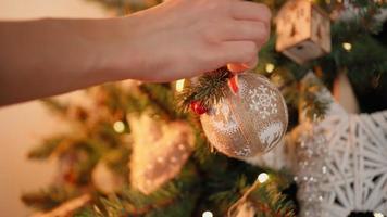 detailopname, een onherkenbaar vrouw siert een Kerstmis boom met feestelijk ballen. warm lichten van slingers flikkeren in de achtergrond. vakantie nieuw jaar. winter atmosfeer, traditie concept. video