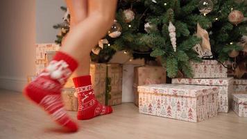 avvicinamento, bellissimo femmina gambe nel rosso nuovo anni calzini siamo adatto per decorazione un' Natale albero su in punta di piedi. nuovo anno, inverno atmosfera, caldo luce. festivo umore. video