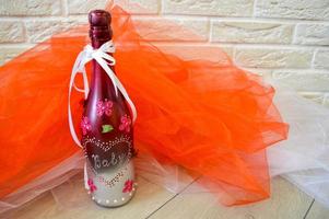 A homemade bottle of champagne decorated with a heart pattern, baby lettering and flowers on a background of red and white fabric and a brick wall. photo