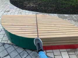 a wooden bench in the center of the residential complex in the form of a droplet. seating area with red and green color. wooden seat for buyers. a bully in a sneaker put his foot on a new bench photo