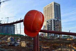 bright orange construction helmet hangs on the fence. against the background of a high multi-storey glass building. there is debris on the helmet and potholes due to old age. protective uniform photo