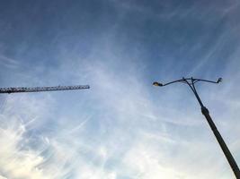 poste de luz con una bombilla de luz blanca contra un cielo despejado. linterna para iluminar dos caminos. pilar gris con una linterna. hay una grúa de construcción cerca. la linterna y la grulla foto