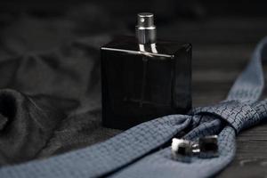 A bottle of mens cologne and cufflinks with blue tie lie on a black luxury fabric background on a wooden table. Mens accessories photo