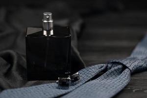 A bottle of mens cologne and cufflinks with blue tie lie on a black luxury fabric background on a wooden table. Mens accessories photo