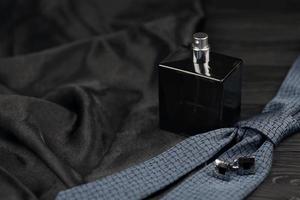 A bottle of mens cologne and cufflinks with blue tie lie on a black luxury fabric background on a wooden table. Mens accessories photo