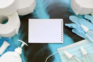 Set of items for Covid-19 quarantine. Empty notepad page and toilet paper, rubber gloves with surgical face mask and hand sanitizer with liquid soap bottle on human chest x-ray film photo