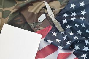 Blank paper lies with knife and army dog tag necklace on camouflage uniform and american flag photo