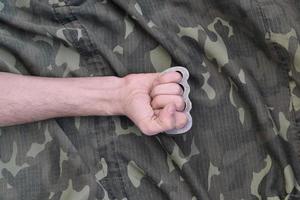 Male fist with brass knuckles on the background of a camouflage jacket. The concept of skinhead culture, handmade melee weapons photo