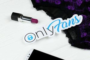 KHARKOV, UKRAINE - FEBRUARY 14, 2021 Onlyfans paper logo, red lipstick with smartphone and black lingerie on white wooden table
