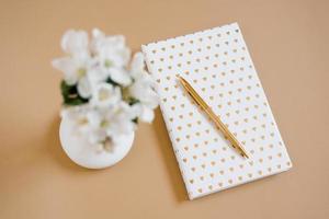 White notebook with gold hearts on the cover, gold pen and white apple flowers in a vase out of focus on a beige table. Layout and design concept. Flat lay. photo