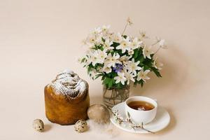 Easter cake craft in. Craft with raisins, candied fruits and a sprinkle of powdered sugar. Homemade cake. Eggs, a cup of tea and spring flowers in a vase on a beige background photo