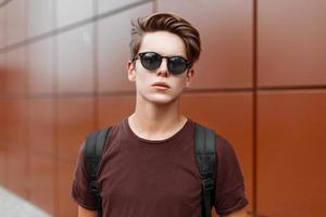 Young handsome guy with hair in sunglasses standing near a wall photo