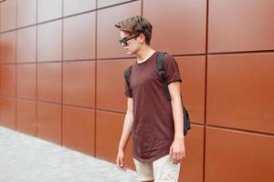 Young handsome guy with a backpack and sunglasses standing near a wall. photo