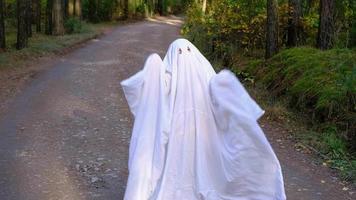 uma criança em lençóis com recortes para olhos como uma fantasia de fantasma dançando em uma floresta de outono assusta e aterroriza. um tipo de fantasma engraçado. festa de halloween, câmera lenta 4k video