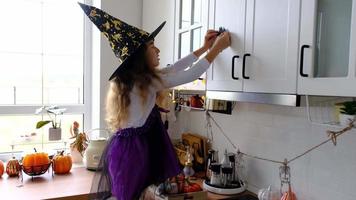 kind siert de keuken in huis voor halloween. meisje in een heks kostuum Toneelstukken met de decor voor de vakantie - vleermuizen, jack lantaarn, pompoenen. herfst comfort in huis, scandi-stijl keuken, zolder video