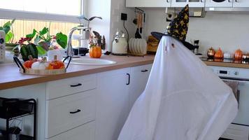 en barn i ark med Skära ut för ögon tycka om en spöke kostym i de kök dekorerad för de halloween Semester. en snäll liten rolig spöke. halloween fest video