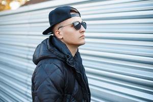 Young handsome man in a black cap and sunglasses in a warm jacket on a background of metal wall photo