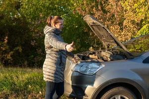 A woman in a jacket is trying to fix a broken car on the road. photo