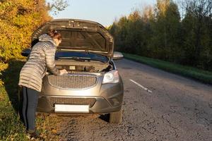 A woman in a jacket is trying to fix a broken car on the road. photo