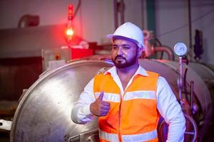 Professional engineer with helmet works to maintain industrial construction equipment. Worker is standing in the factory with thump up. photo