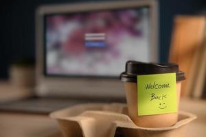 volver al concepto de trabajo. primer plano de la nota de bienvenida en la taza de café para llevar en el escritorio de la oficina. mensaje de un colega o jefe foto