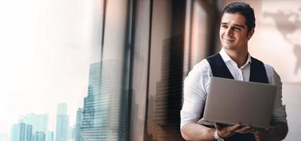 Portrait of a Young Confident CEO or Leader Working on Computer Laptop in the Modern Workplace. Smiling Businessman Standing by the Window and Looking outside photo