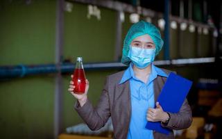 trabajador científico en una fábrica de bebidas embotelladas con sombrero de seguridad posando para comprobar la calidad de los productos de semillas de albahaca en la cinta transportadora antes de su distribución al negocio del mercado. foto