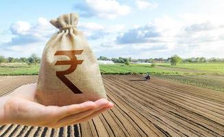 Hand holds out a indian rupee a farm field. Financial support, subsidies for agriculture. Farm loans, economic preferences. Purchase or lease rent of land. Payment of taxes for use natural resources. photo