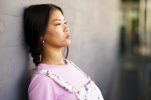 Young Chinese woman with a lost look and a serious expression leaning against an urban wall. photo